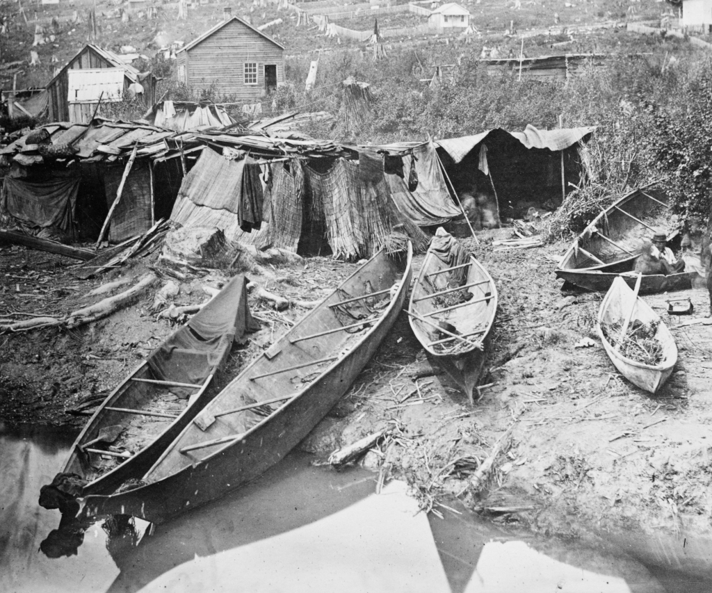 Historical photograph of wooden canoes on a muddy riverbank with wood stick-frame structures in background.