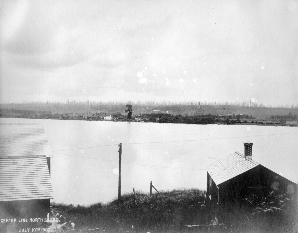 Historical photograph of a forested riverbank scattered with low buildings and a prominent church.