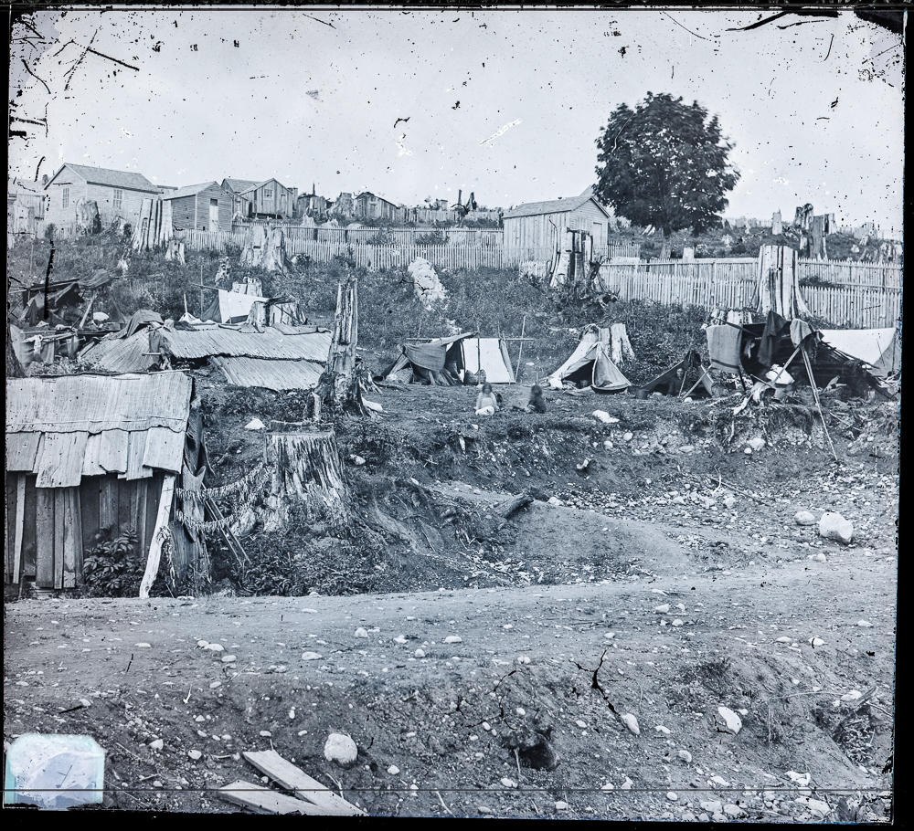 Historical image of simple wooden buildings and fences on a hill, with a string of dead fish hung between a stump and a building.